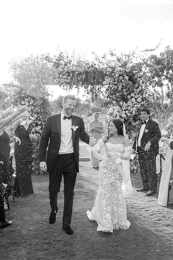 Wedding recessional as newlyweds walk hand in hand down the outdoor aisle under a floral arch, confetti falling on veil and tuxedo