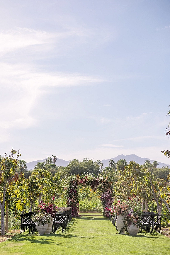 Ceremony setup for an outdoor wedding ceremony with a floral arch accent, green aisle runner, black chairs, and mountains beyond palm trees