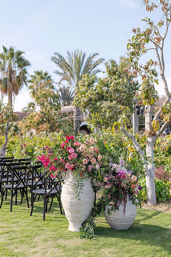 Ceremony aisle florals with wedding aisle urns, lush roses and trailing greenery lining black chairs on a sunny garden lawn by palm trees