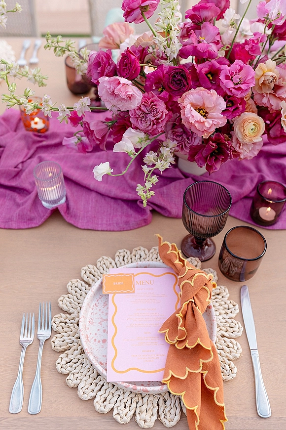 Reception tablescape with wedding table centerpiece of pink roses, ranunculus, and sweet peas on a wood table with candles and colored goblets