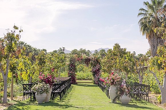 Ceremony setup with a floral arch and aisle arrangements, wooden chairs lining a grass aisle beneath orchard trees with mountains beyond