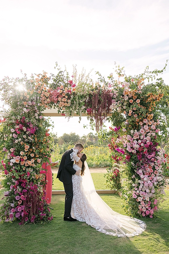 Wedding kiss as the couple kissing ceremony beneath a rose-and-greenery floral arch with hanging amaranthus on a sunlit garden lawn