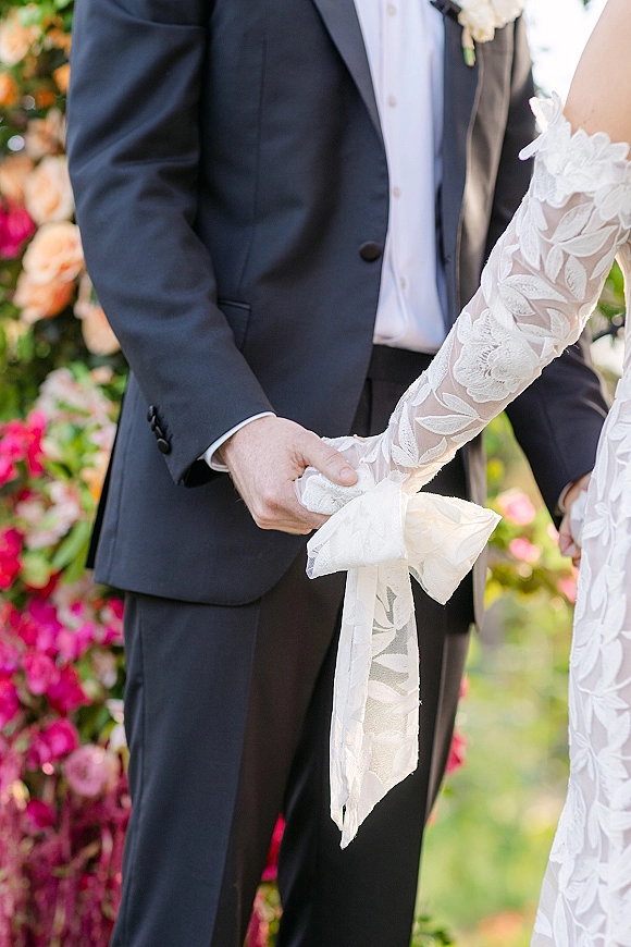 Ceremony moment during a handfasting ceremony as bride in lace sleeves and groom in suit hold hands with a cord under a floral arch