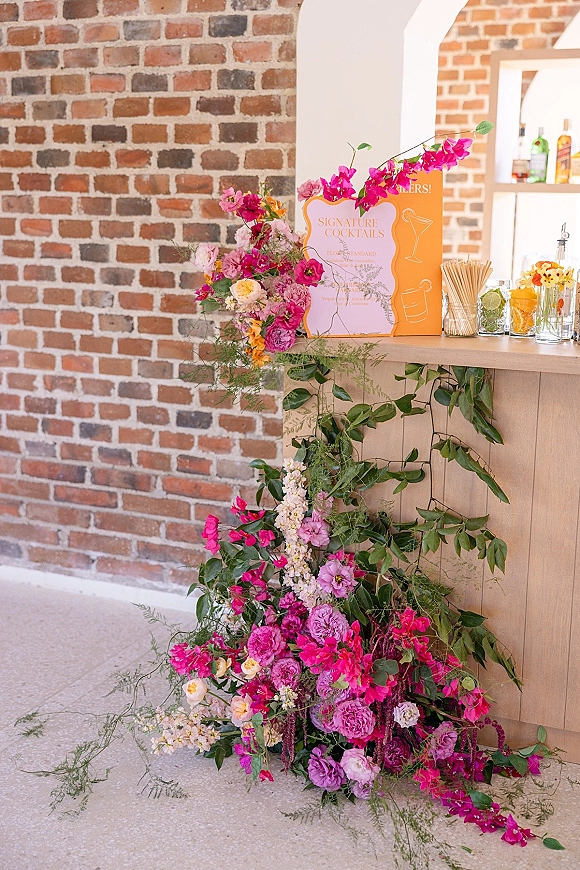 Cocktail bar decor with a signature cocktails sign, hot pink florals and greenery garland, glassware and bottles against a brick wall backdrop
