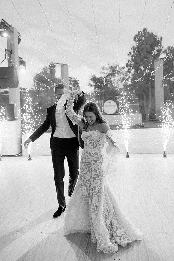 First dance moment with bride in lace wedding dress twirling with groom in suit under string lights and cold spark fountains outdoors