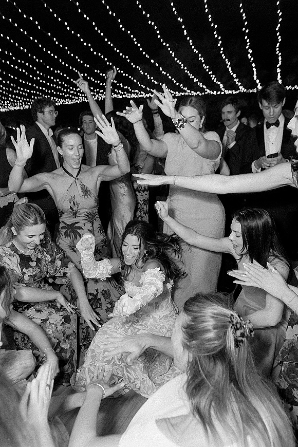 Wedding dance floor packed with guests dancing in a lively circle under string lights at an outdoor nighttime reception, hands in the air