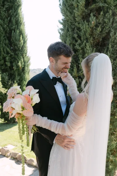 Couple portrait of bride touching groom’s face as he laughs in a close embrace, holding a calla lily bouquet on a stone path by evergreens