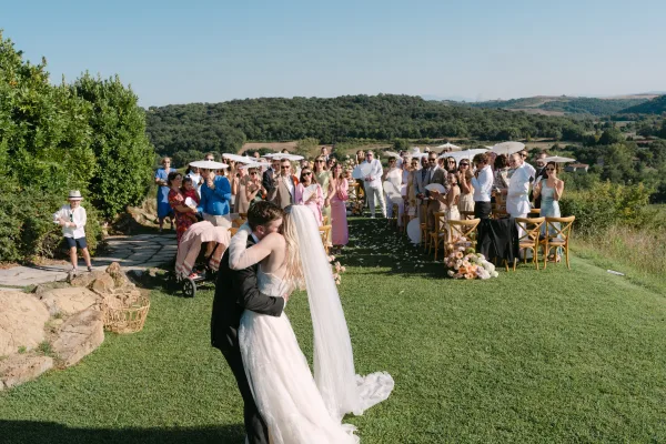 Wedding kiss as the bride and groom embrace on a stone path, veil trailing, guests with white parasols on a sunny hillside lawn ceremony