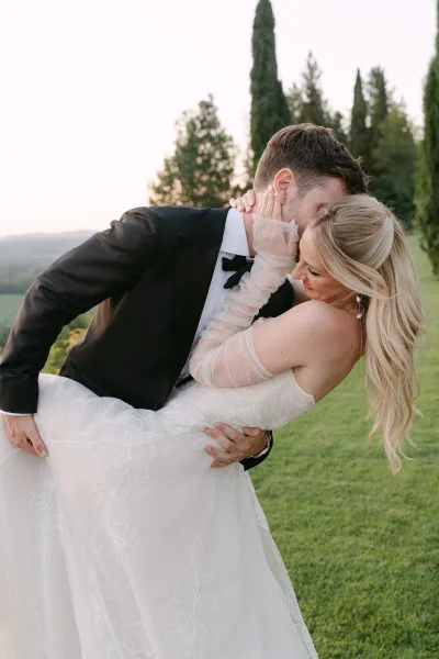 Wedding kiss portrait of groom dipping the bride in a black tuxedo, her lace gloves on his face, on a green lawn with trees and hills