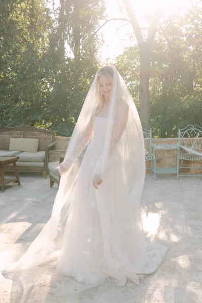 Bridal portrait of a bride looking down in a lace wedding dress with cathedral veil and long train on a sunlit patio by trees