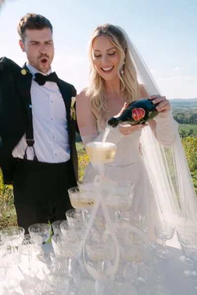 Champagne tower moment as bride and groom pour bubbly into stacked coupe glasses, veil and tuxedo in sunny hills backdrop