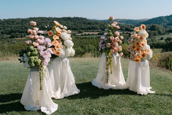 Ceremony floral arrangements with peach and blush roses, gerbera daisies and hydrangeas on draped pedestals against rolling hills and blue sky