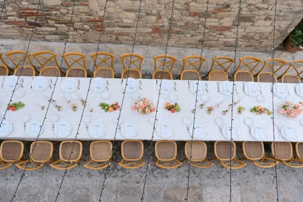 Reception tablescape on a long wedding reception table with white linens, cross-back chairs, taper candles, florals, fruit, and string lights by a stone wall