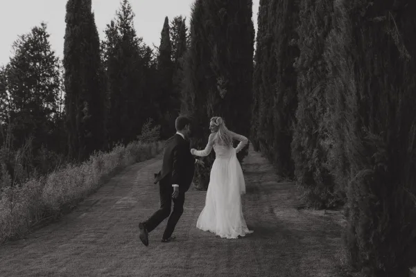 Couple portrait in a black and white wedding photo, bride and groom walking away hand in hand down a tree-lined path, veil trailing