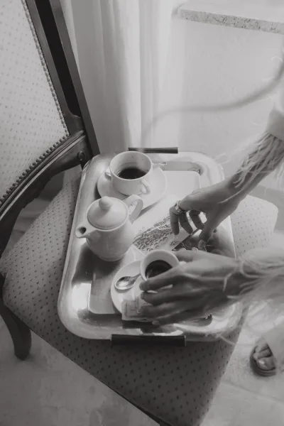 Bridal jewelry details with a wedding ring flatlay beside a coffee cup on a tea tray, set near an upholstered chair and window curtain