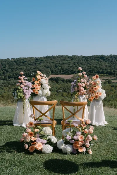 Ceremony altar decor with pastel floral ceremony pillars and draped fabric beside crossback chairs on a hillside lawn under blue sky