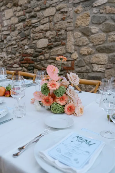 Reception tablescape with a wedding table centerpiece of peach and pink flowers, roses, and fruit on white linen beside a stone wall