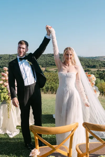 Recessional moment as newlyweds raising hands walk up the aisle, bride in long veil and gloves beside groom in tuxedo on a sunny lawn with hills