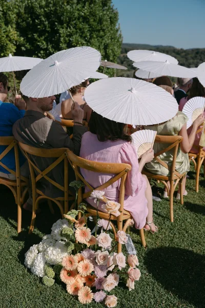 Wedding ceremony guests seated on wooden crossback chairs, many holding white parasols, on a sunny lawn with trees and hills beyond