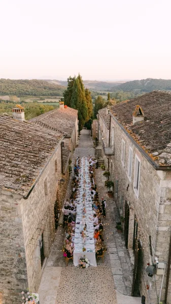 Outdoor reception tablescape with a long banquet table wedding setup, candles and florals under string lights in a cobblestone alley between stone buildings