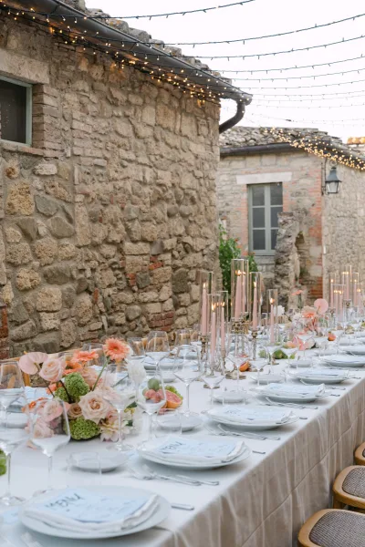 Reception tablescape with taper candles, blush and coral florals, and fruit on a long banquet table under string lights in a stone courtyard