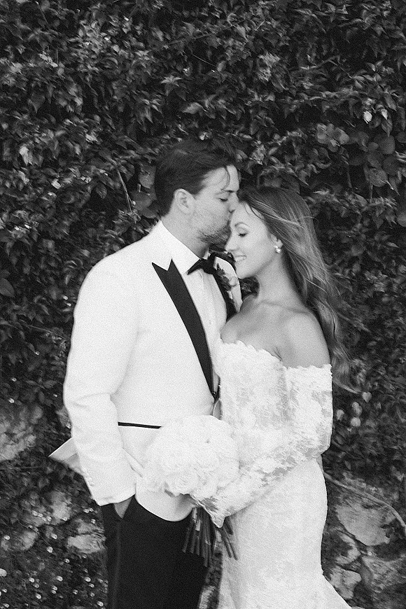 Couple portrait in black and white wedding portrait style, bride and groom kissing as he kisses her forehead before a leafy hedge backdrop