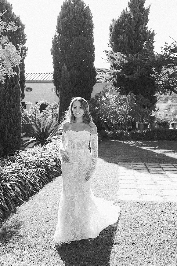 Bridal portrait of a bride in a lace wedding dress with long sleeves and train, standing on a stone path in a garden by a villa