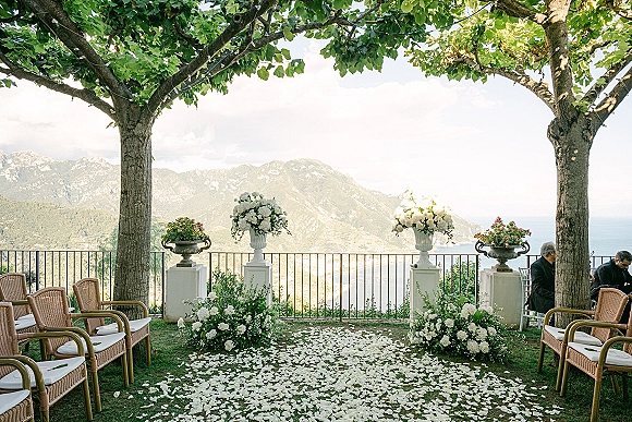 Ceremony setup with outdoor wedding ceremony seating, white florals in urns and a rose petal aisle on a terrace overlooking ocean and mountains