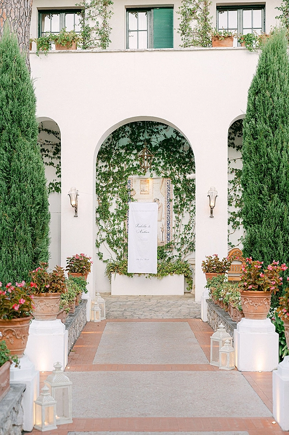 Wedding welcome sign on hanging fabric with calligraphy, flanked by white lanterns and candles at an arched stucco courtyard entryway