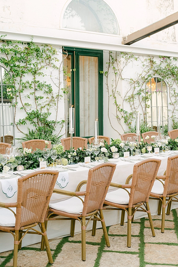 Reception tablescape with greenery garland runner, white roses and hydrangea, taper candles in glass holders, set on a stone patio by vine wall.