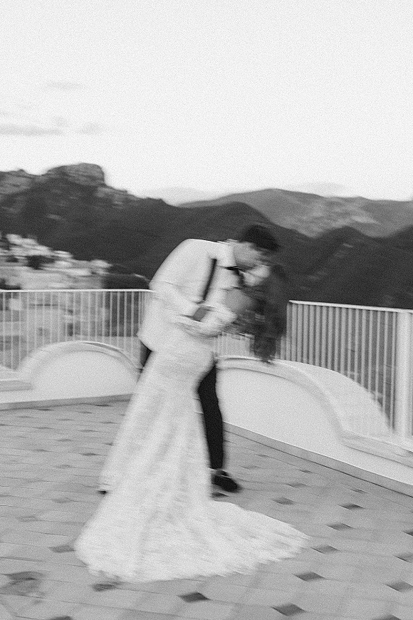 Wedding kiss portrait of groom lifting bride in a dip, her lace gown train flowing as she holds a bouquet on a mountain terrace overlook