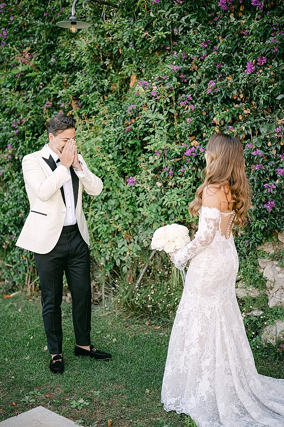 Wedding first look as the groom emotional reaction shows him covering his face while the bride approaches with a white rose bouquet by a hedge and stone wall