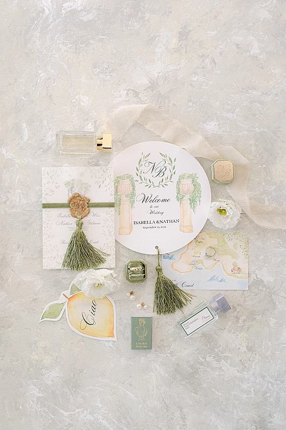 Invitation suite wedding invitation flat lay with wax seal, green tassels, rings and perfume on a light gray textured surface with sheer fabric