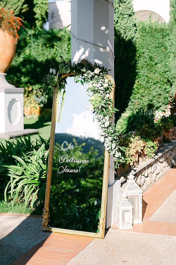 Wedding welcome sign on an ornate gold mirror with calligraphy lettering, greenery garland and white flowers on a garden patio with lanterns