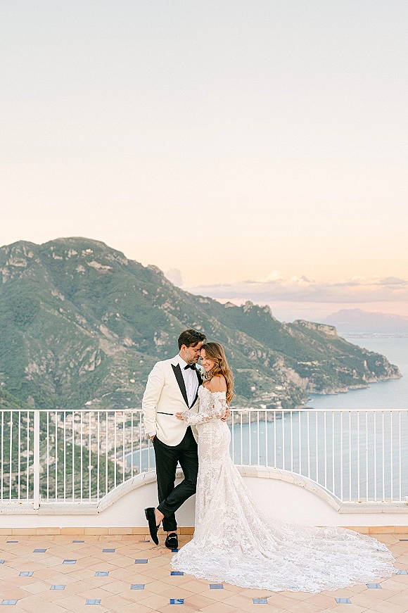 Couple portrait of bride and groom embrace, her lace gown with long train and his white tuxedo, on a terrace with ocean and mountains behind