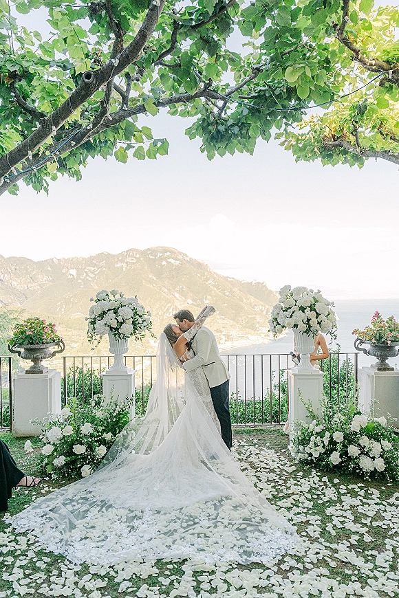 Wedding kiss as bride in lace dress and cathedral veil meets groom in suit on ocean-view terrace aisle with white rose urns and petals