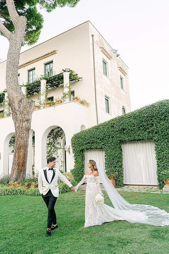 Couple portrait of bride and groom holding hands, her long veil and bouquet flowing, beside ivy-covered villa arches and curtains