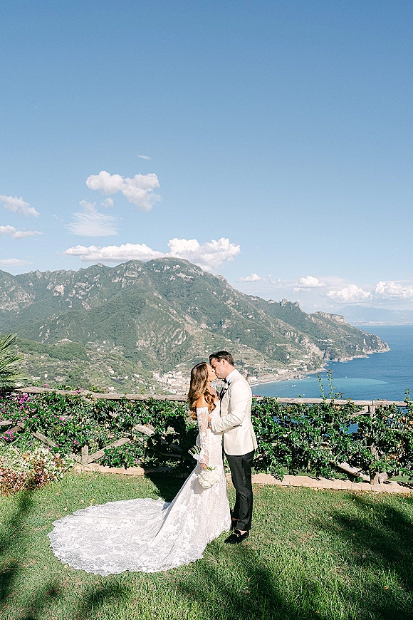 Wedding kiss portrait of bride and groom kissing, her lace gown train and bouquet against a mountain-and-ocean view under blue sky