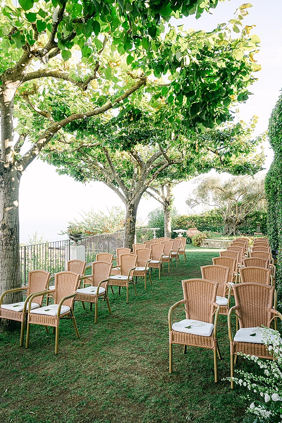 Ceremony setup with outdoor wedding ceremony seating of wicker chairs and white cushions lining a floral aisle on grass with ocean view