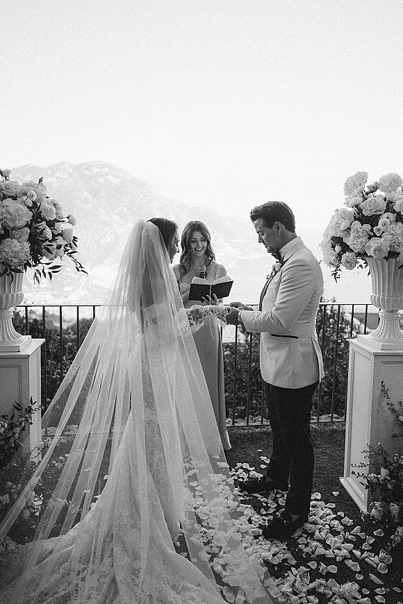 Wedding vows as bride and groom hold hands on a terrace, long veil trailing by rose petals, with mountain view behind