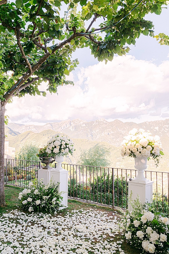 Ceremony aisle decor with a white flower petal aisle, lined by white rose urns on pedestals beneath trees with mountain views