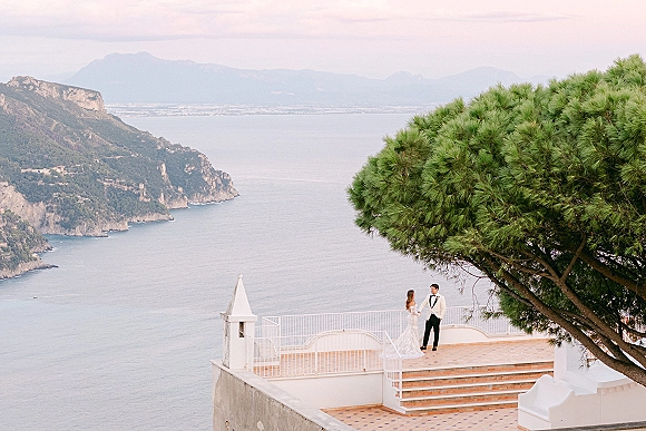 Couple portrait on stone steps, bride in lace wedding dress and veil holding a bouquet beside groom in tuxedo, ocean cliffs behind
