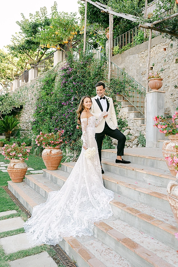 Couple portrait of bride and groom on stairs, bride in lace sleeve gown with long train and white bouquet, groom in tux by stone wall greenery