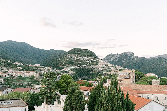 Mountain town landscape with terracotta roof tiles and hillside church buildings, cypress trees, and forested hills beneath a cloudy sky