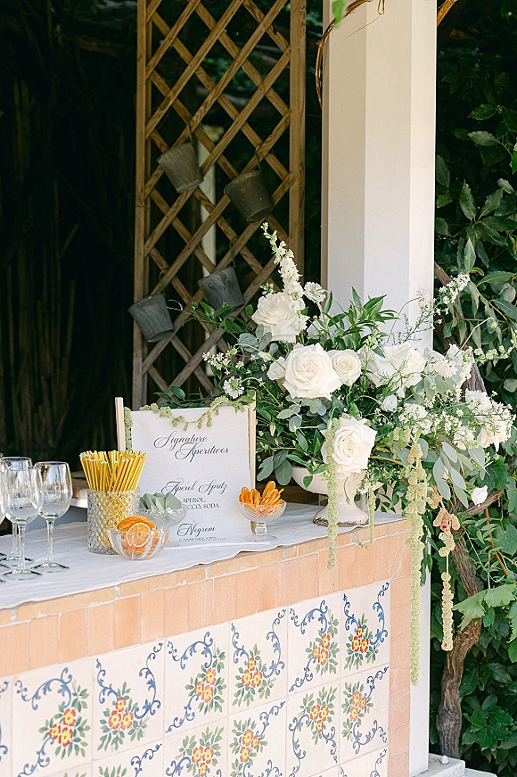 Wedding bar setup with cocktail station wedding menu sign, white roses and greenery, gold stirrers, and wine glasses against garden trellis backdrop