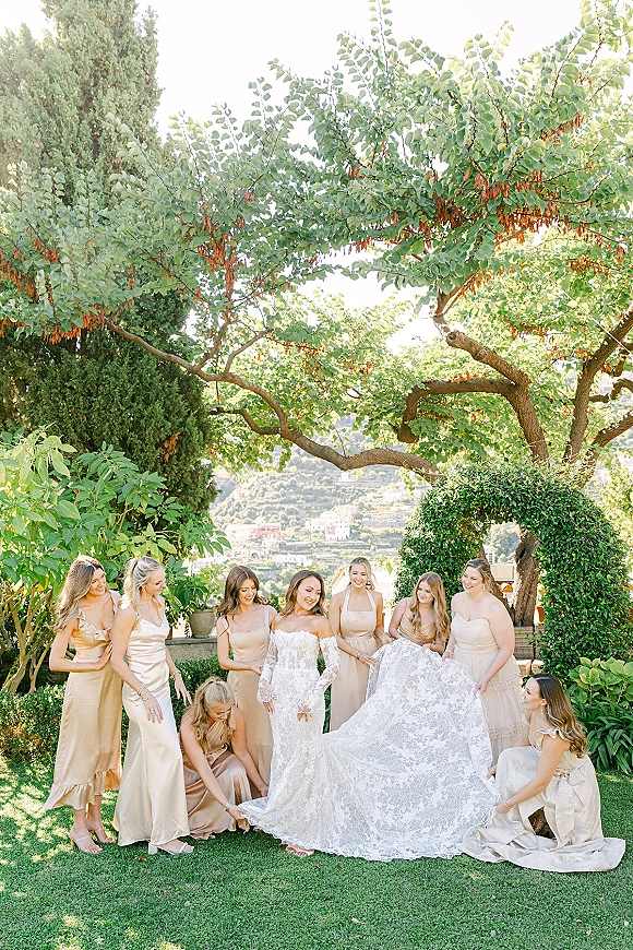 Bride with bridesmaids gathered around her lace gown, holding the dress train and laughing on a garden lawn by a stone terrace wall