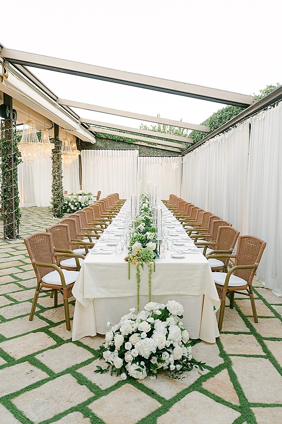 Reception tablescape with a long banquet table wedding setup, ivory linens, white rose garland runner, taper candles, and chandeliers on a covered patio
