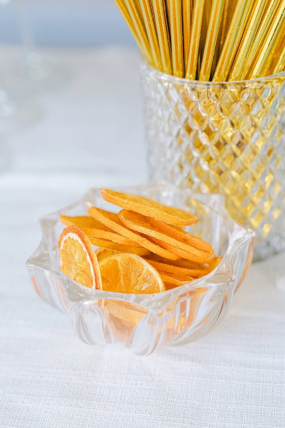 Wedding drink garnishes arranged with dried orange cocktail garnish, gold stirrers, and crystal bowls on a white linen tabletop in soft light