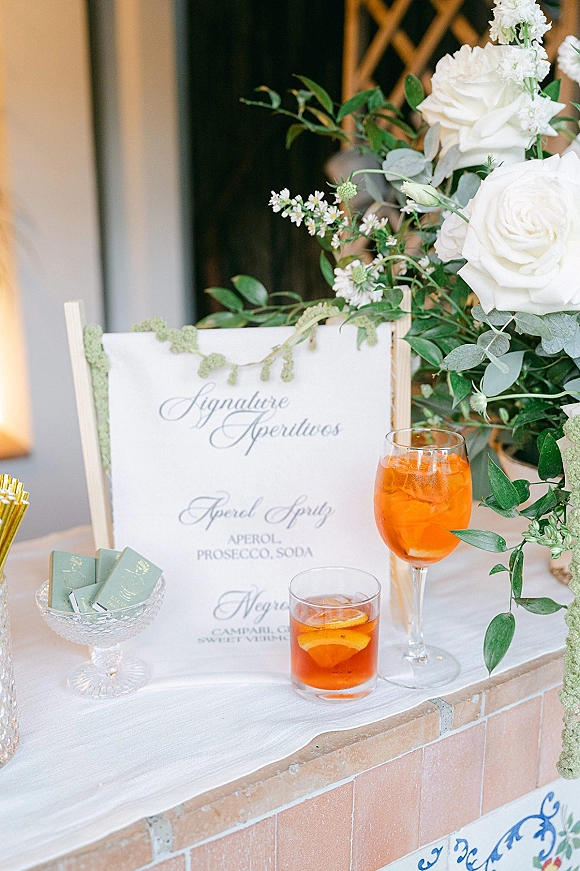 Signature drinks sign with wedding signature cocktails in orange glasses, framed in wood with white roses and greenery on a tiled bar counter