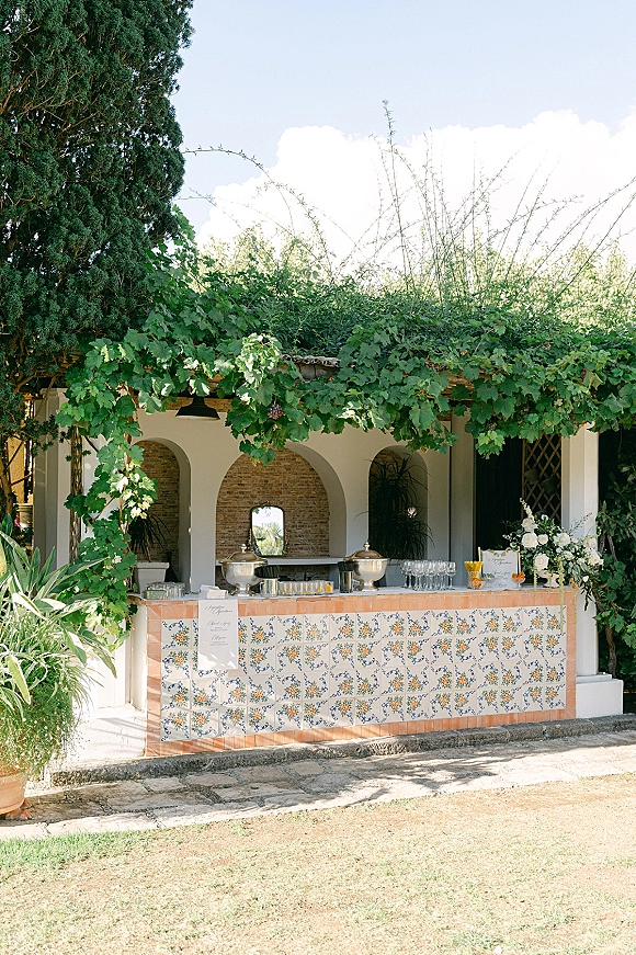 Wedding bar setup with patterned tile front, glassware and drink dispensers under an ivy-covered pergola with stucco arches behind